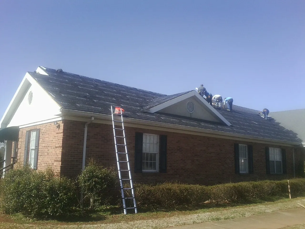 Crew tearing shingles off a brick ranch roof with a ladder set — Emergency Roof Repair work by our crew in Islip Terrace