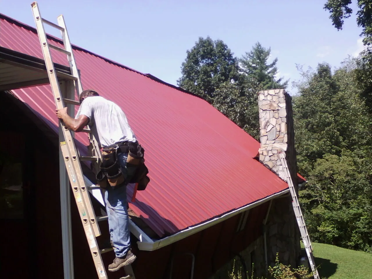 Roofer on a ladder beside a red metal roof and stone chimney — Soffit & Fascia Repair by Roofing Brothers in Islip Terrace
