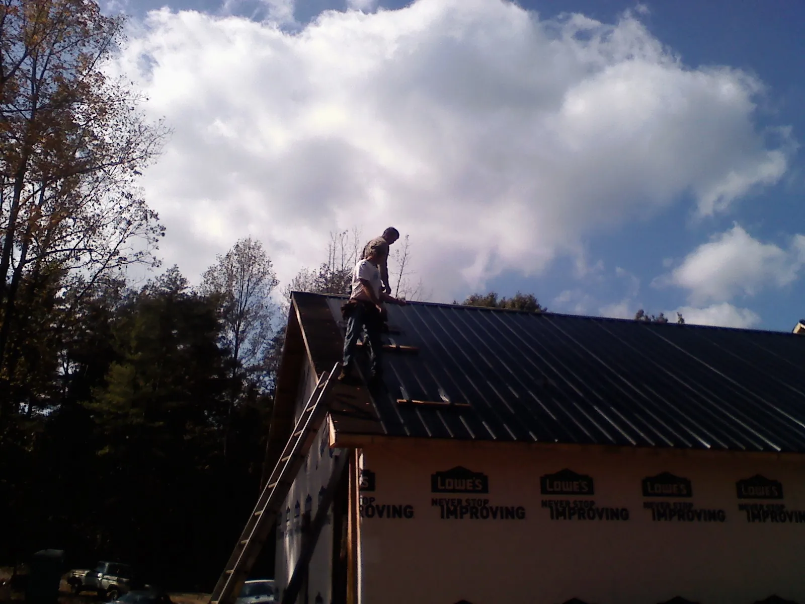 Roofer fastening dark metal panels high against a cloudy sky, from a Roof Inspection job in Islip Terrace